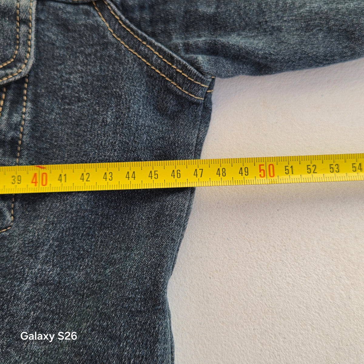 Mesure de la largeur de la veste en jean More & More taille 40 avec un mètre ruban jaune.