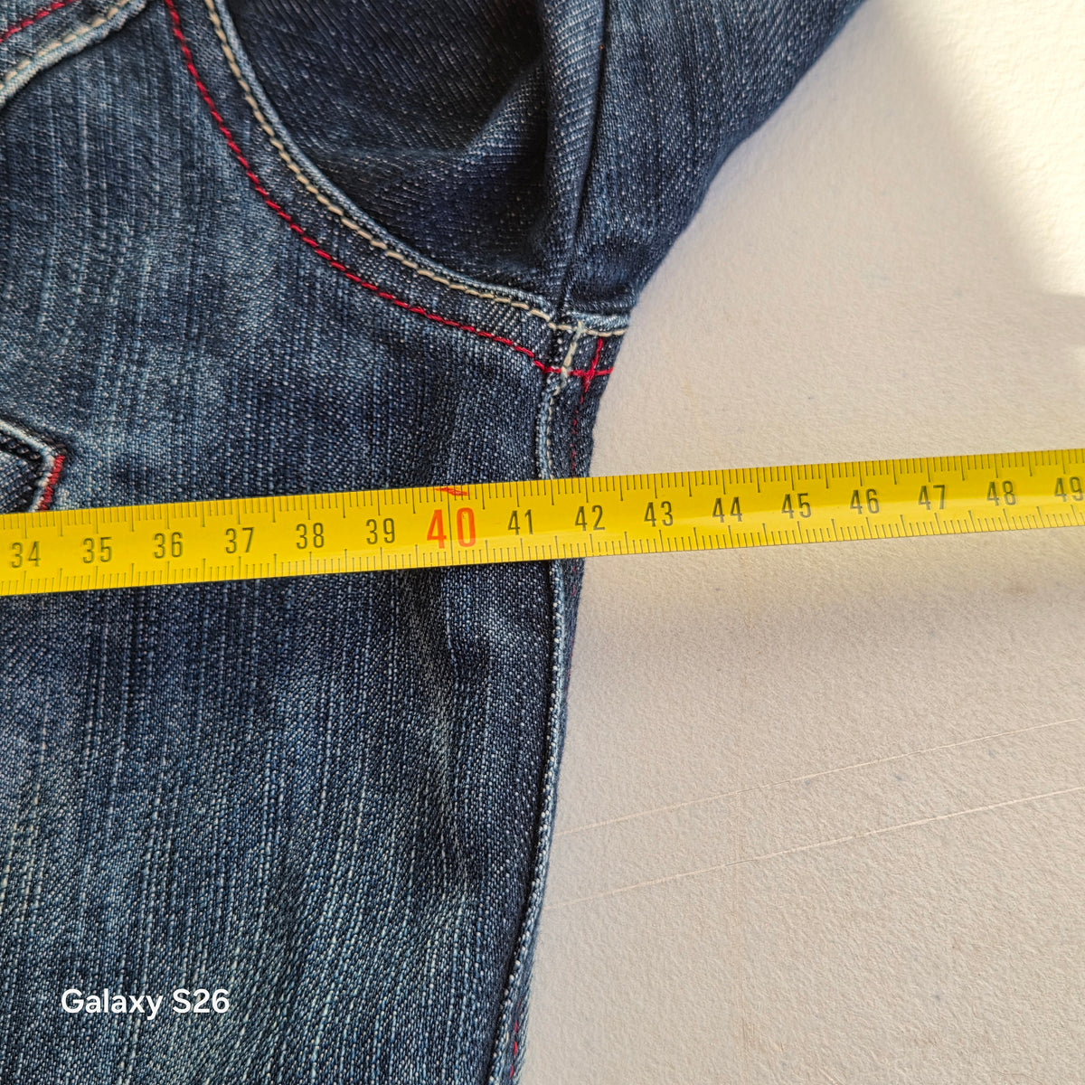 Mesure de la largeur de la veste en jean Naf Naf taille S avec un mètre ruban jaune.